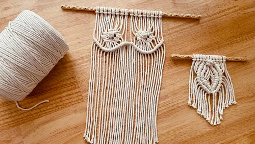 Macrame wall hangings hang next to a spool of thread on a wooden surface.