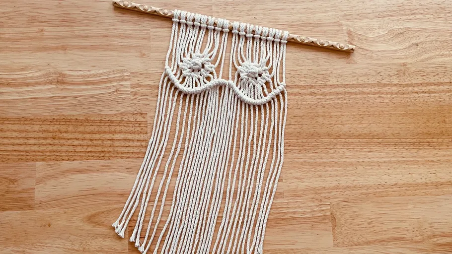 Macrame wall hanging on a wooden stick over a wooden background.