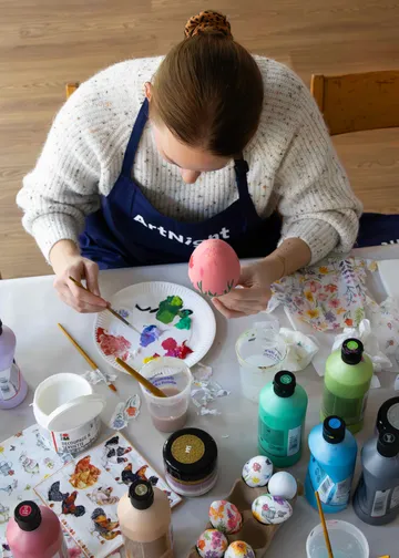 Person paints egg surrounded by art supplies.