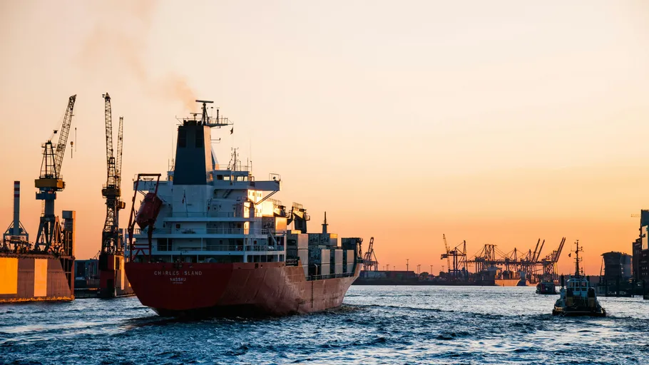 Cargo ship sailing in industrial port at sunset.