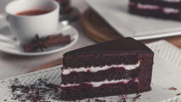 Chocolate cake slice on plate, tea in background.