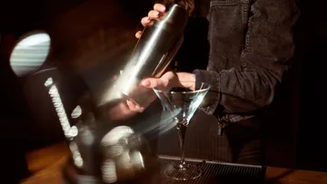 Barkeeper schüttelt Cocktailmixer, Glas daneben.