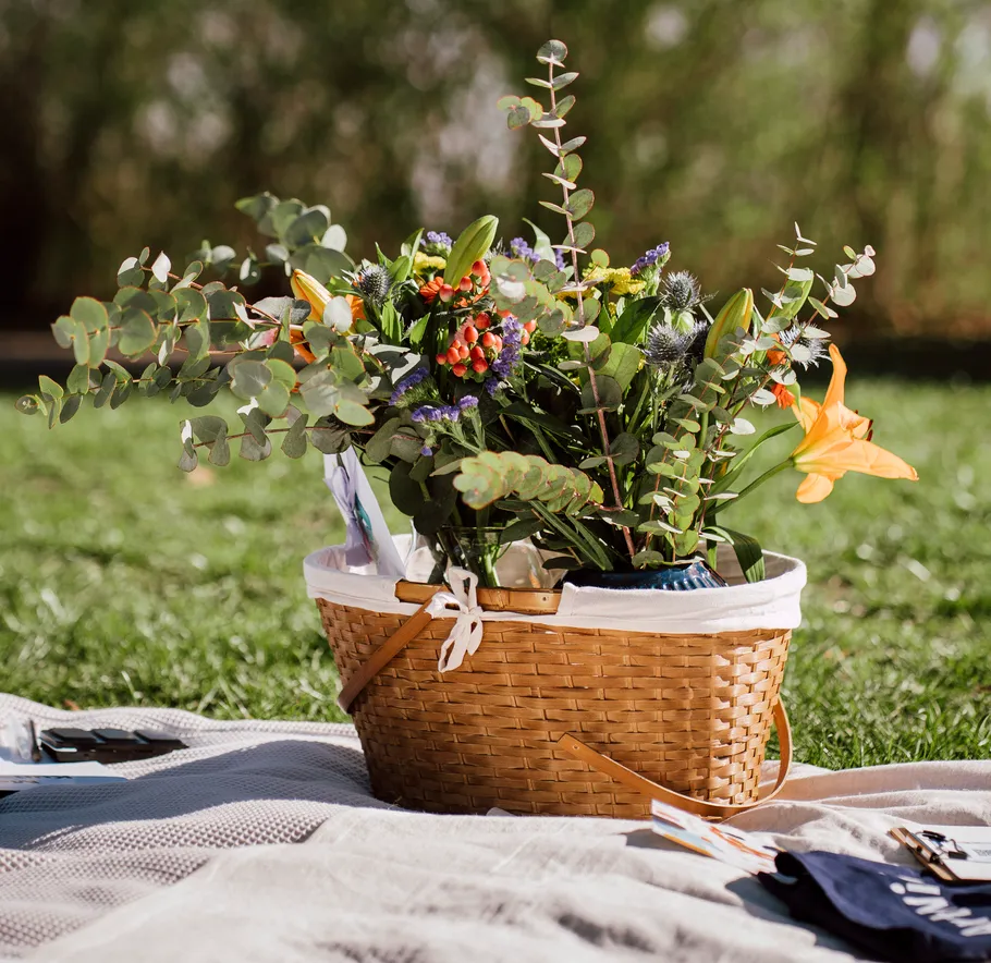Flower-filled basket on picnic blanket outdoors.