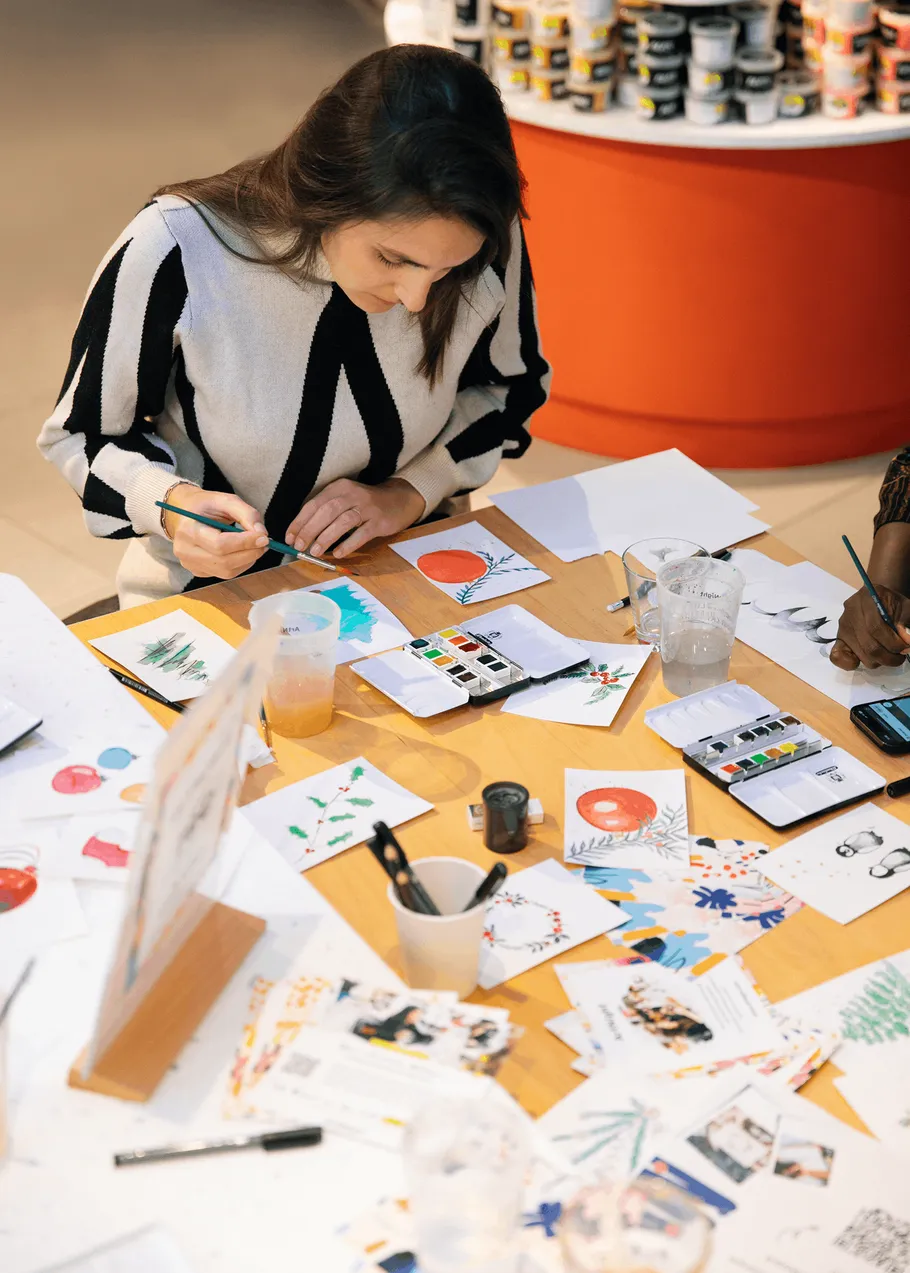 Woman painting at wooden table, art supplies around.