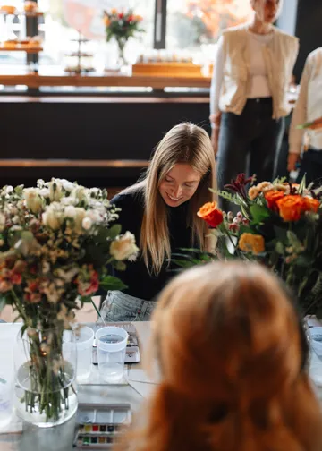 Woman arranging flowers indoors with others nearby.