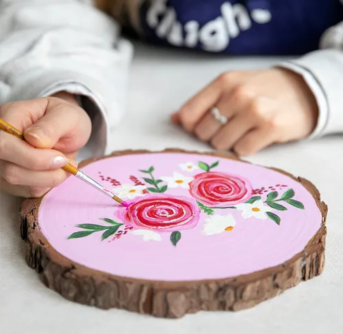 Person painting flowers on wood slice indoors.