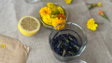 Glass with blue flowers, lemon, and marigolds nearby.