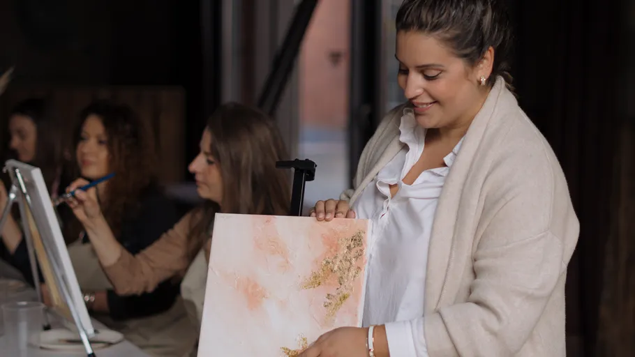 Woman holds painting, other women paint nearby.