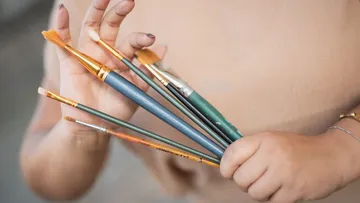 Hand holding various paintbrushes, blurred background.