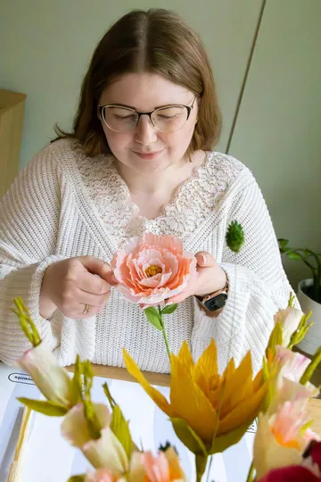 Frau bastelt Papierblume an einem Tisch.