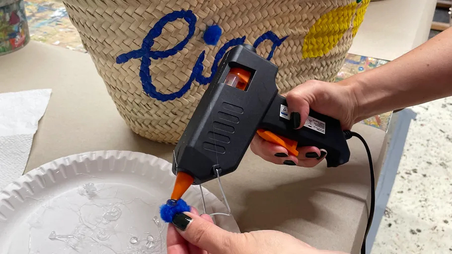 Person gluing pom-pom on straw bag.