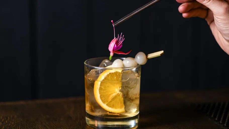 Hand garnishing cocktail with flower on table.