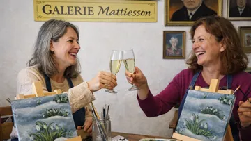 Two women toast with drinks painting in gallery.