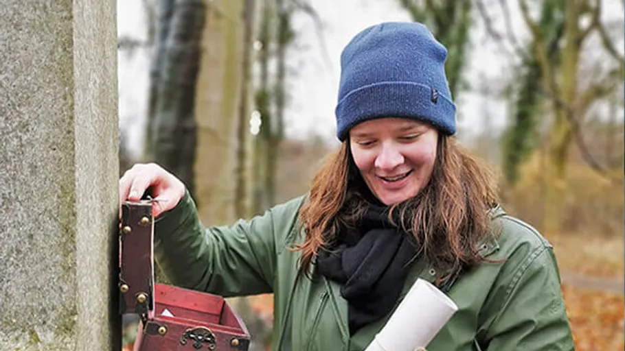 Person öffnet Kiste, hält Schriftrolle im Wald.