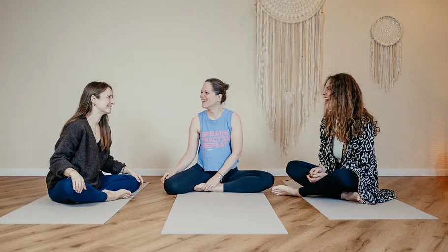 Drei Frauen sitzen drinnen auf Yogamatten.