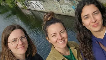 Drei Frauen lächeln nahe einem Graffiti-bedeckten Kanal.