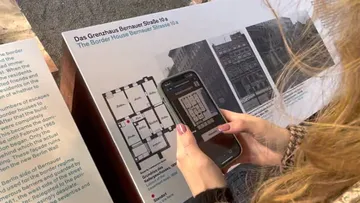 Person fotografiert historische Informationstafel mit Handy.