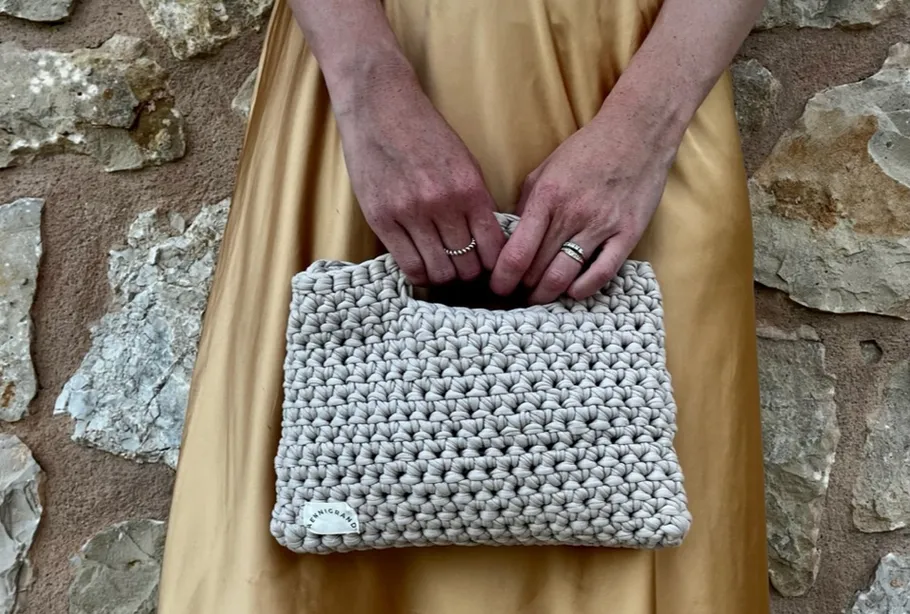 Frau hält gehäkelte Tasche vor Steinwand.