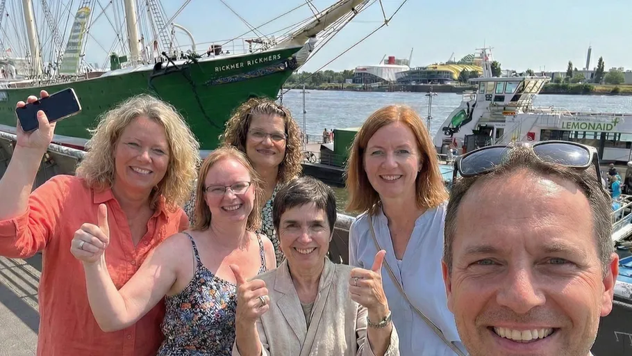 Group smiling near ship in sunny harbor.