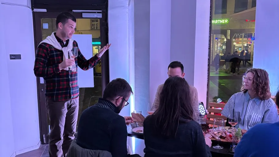 Man standing speaks to seated people indoors.