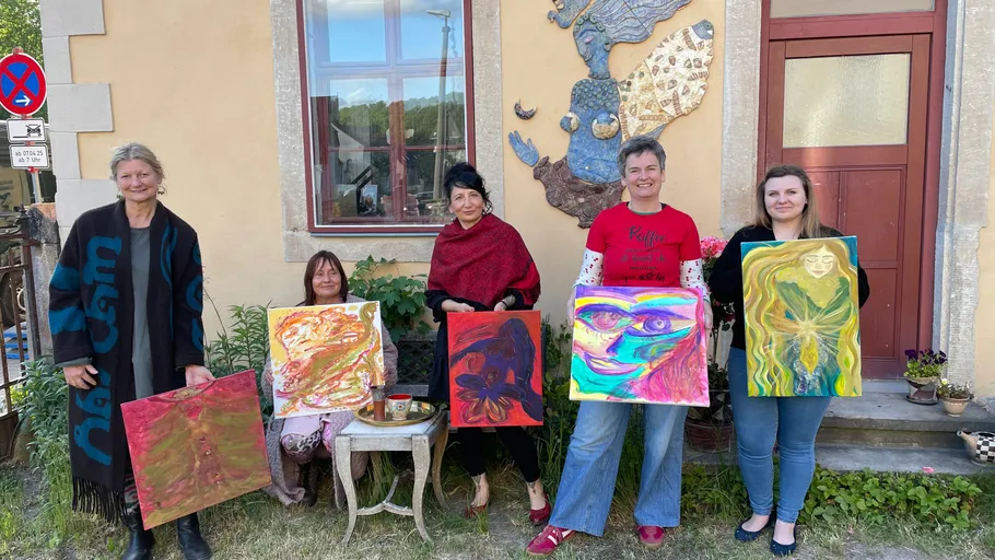 Five women displaying colorful paintings outdoors.