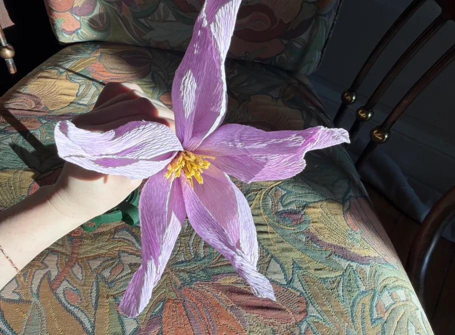 Hand holding purple crepe paper flower indoors.