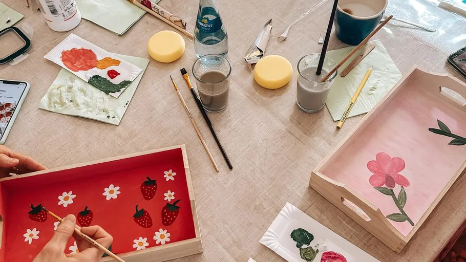 People painting floral designs on wooden trays.