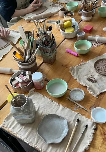 Kunstmaterialien auf Holztisch, Person formt Ton.