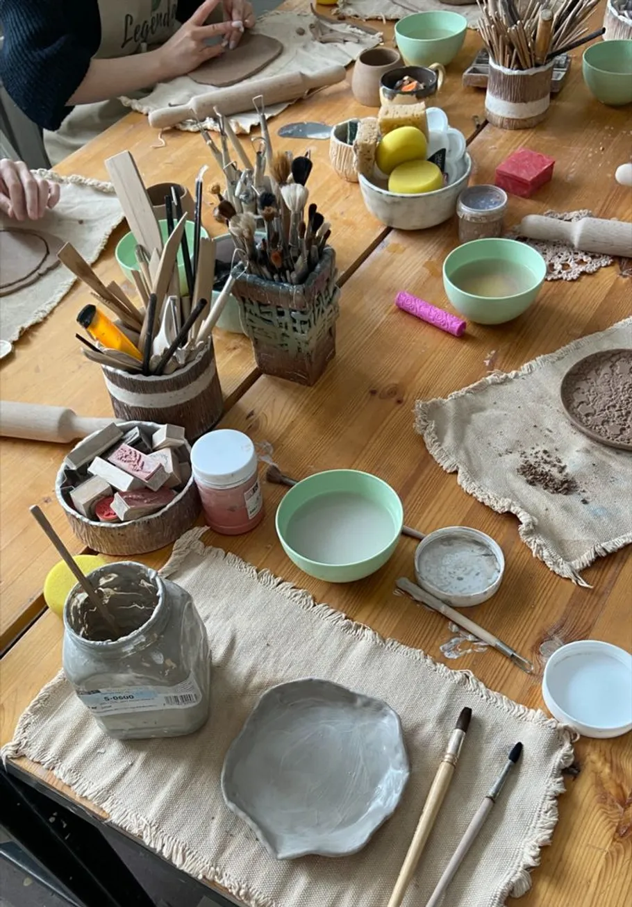 Kunstmaterialien auf Holztisch, Person formt Ton.