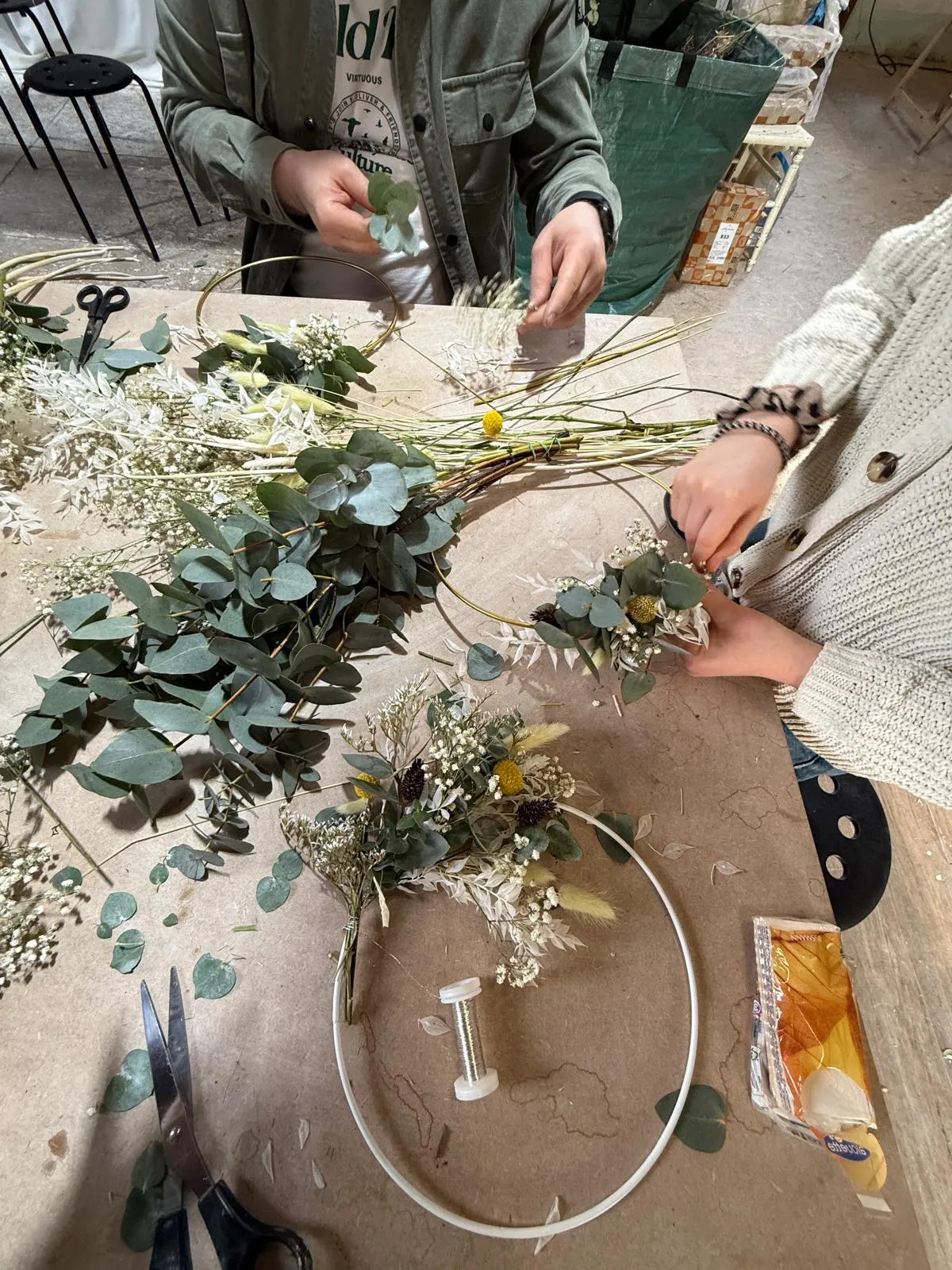 Two people making floral wreath on table.