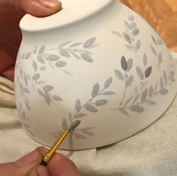 Person painting floral design on ceramic bowl.