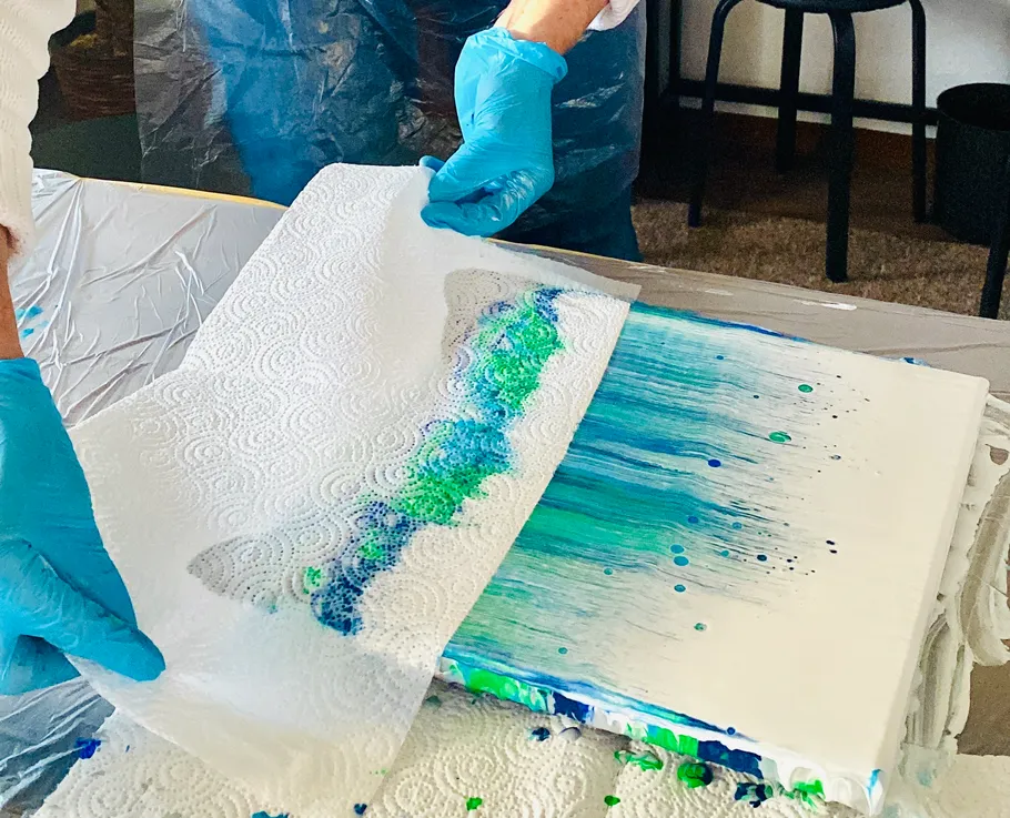 Hands lift paper towel off painted canvas.