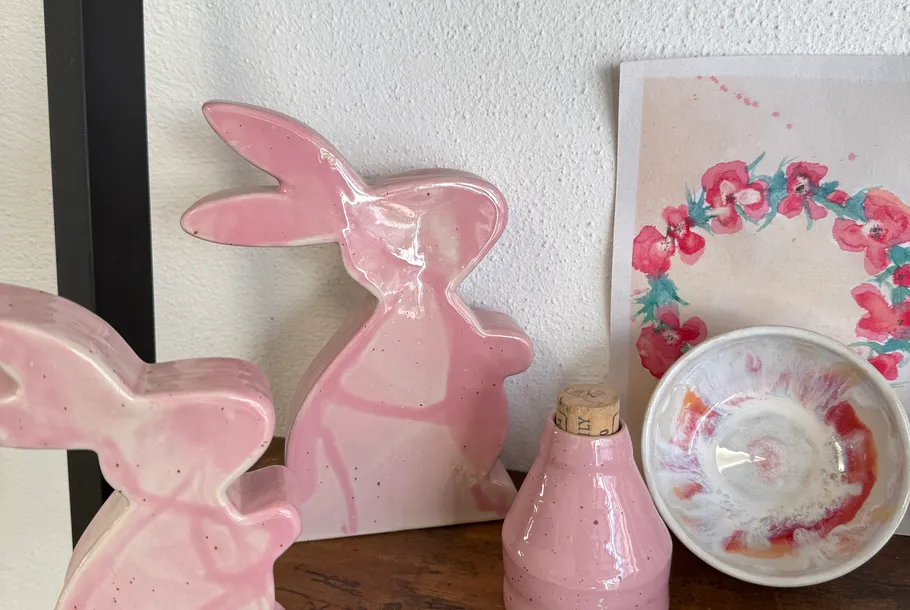 Pink ceramic rabbits and art on wooden shelf.