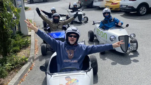 Five smiling people driving mini cars outdoors.