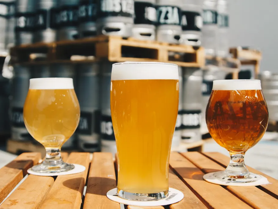 Drei Biergläser stehen auf einem Holztisch vor Fässern.