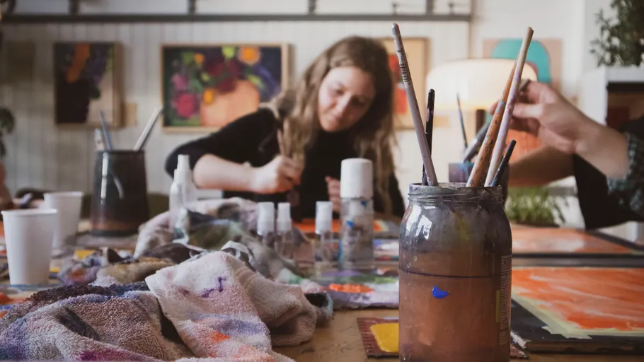 Frau malt an vollgestelltem Kunsttisch drinnen.