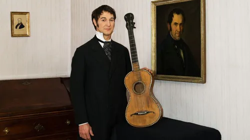 Man in suit holding guitar in room with paintings.