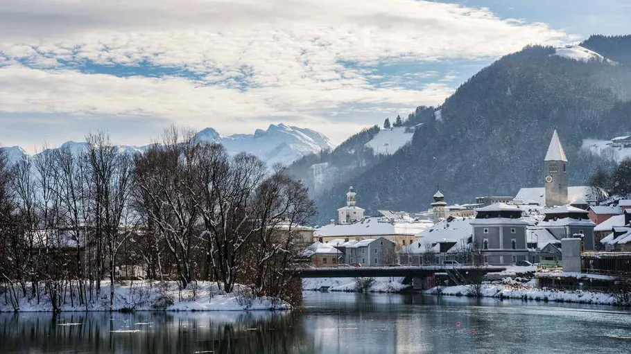 Snowy town by river with mountain backdrop.