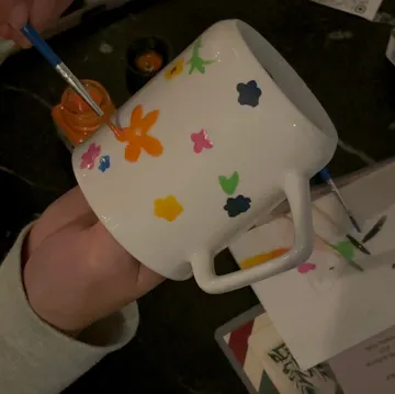 Person malt bunte Blumen auf weiße Tasse.