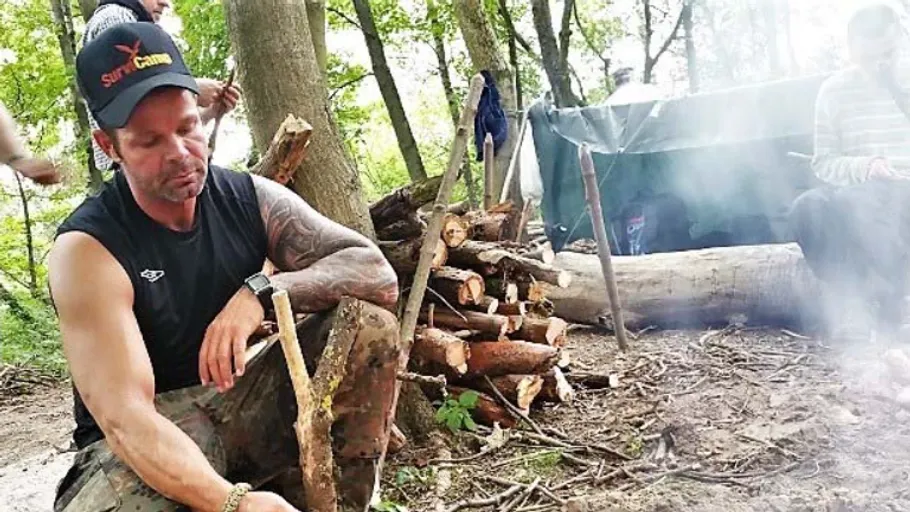 Mann im Wald sitzt bei Holzhaufen und Zelt.