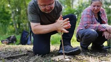 Mann macht Feuer mit Stock, Frau schaut zu, draußen.
