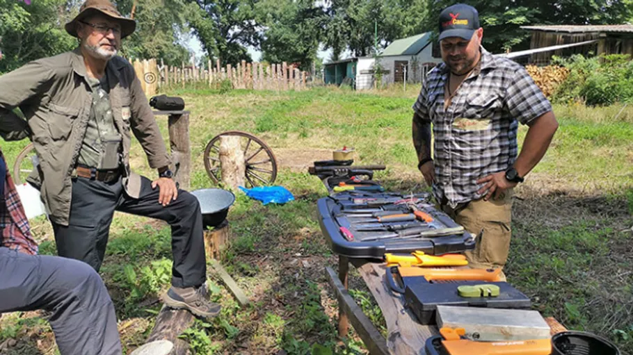 Two men examining knives on outdoor table.