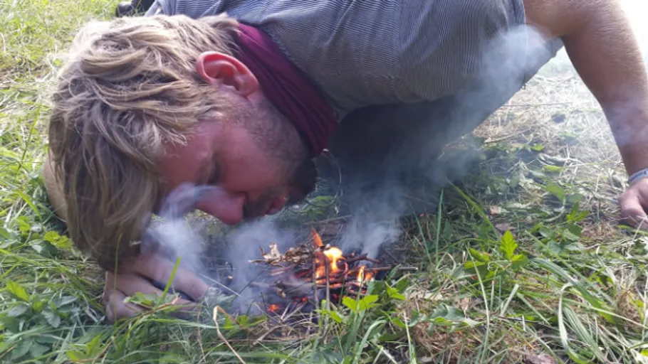 Man blowing on small fire in grassy area.