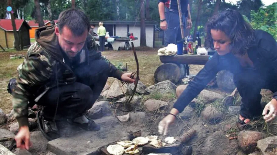 Two people cook on campfire in forest.