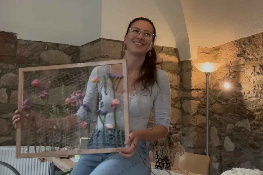 Frau hält Blumenrahmen drinnen, lächelt.