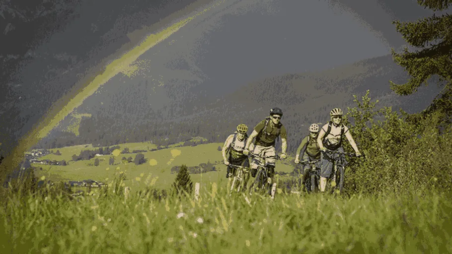 Cyclists biking uphill under a rainbow, forested area.