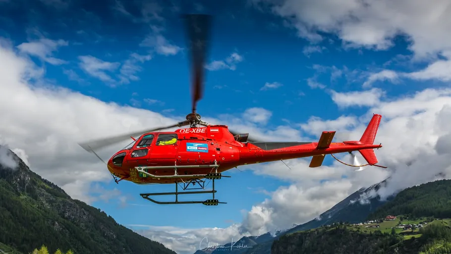 Red helicopter flying over mountainous landscape.