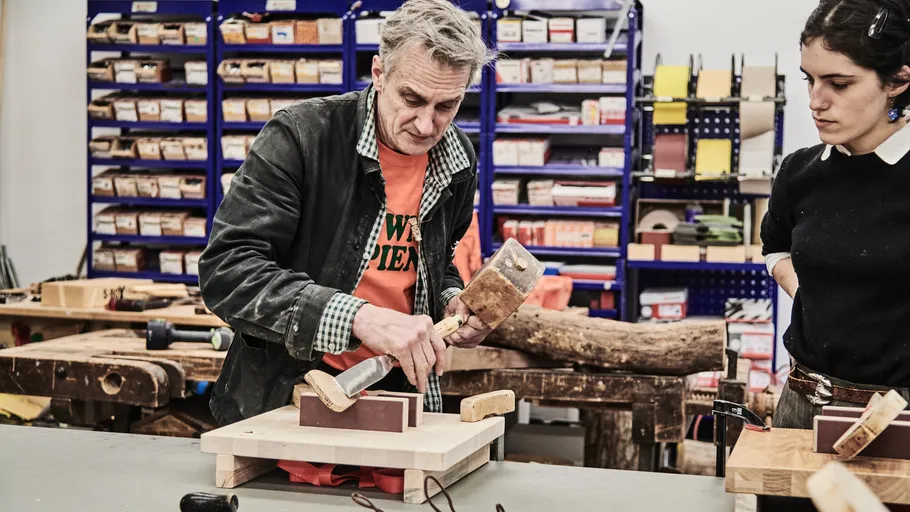 Man demonstrating woodworking technique to woman, workshop setting.
