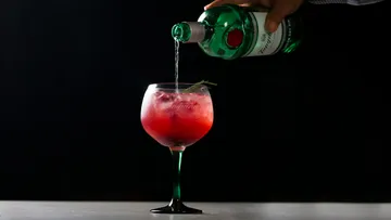 Grüne Flasche gießt Getränk in rotes Cocktailglas.