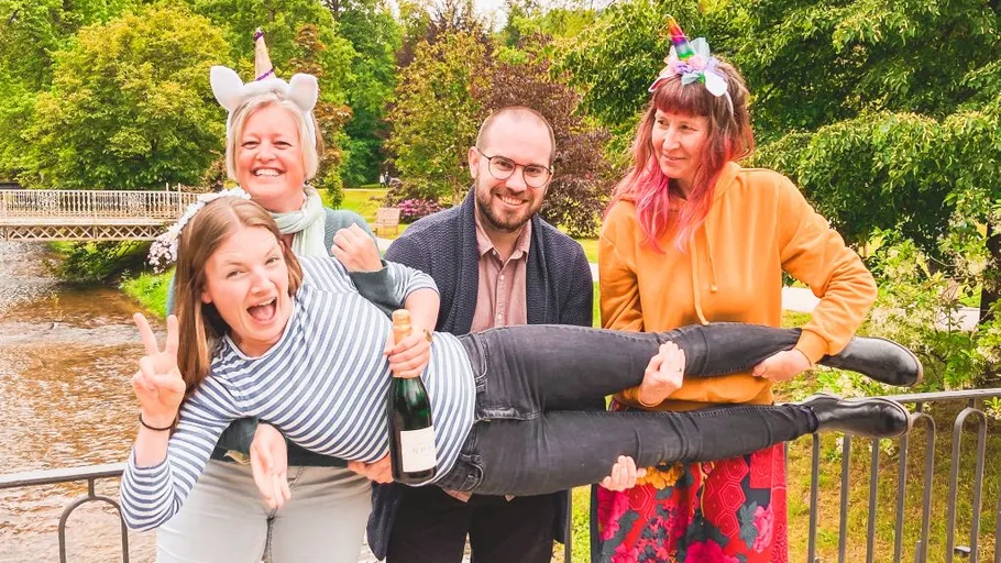 Three people hold a smiling person by a river.